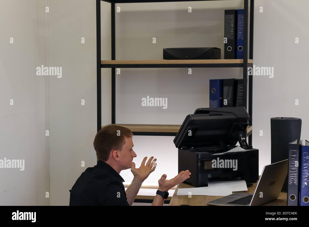 Giovane uomo in camicia nera inginocchiato nella disperazione, mani alzate verso una stampante da ufficio malfunzionante con carta inceppata sulla scrivania. Foto Stock