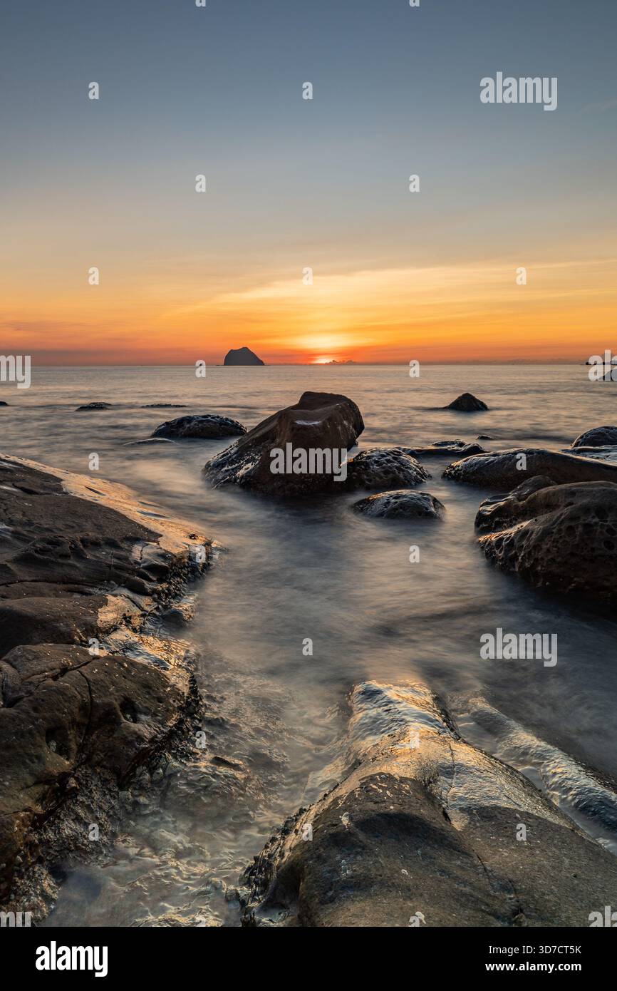 Keelung Waimushan God Table, Taiwan. Il crepuscolo bacia la costa rocciosa mentre le onde nebbiose e le luci dorate tracciano un tranquillo percorso verso l'orizzonte lontano. Foto Stock