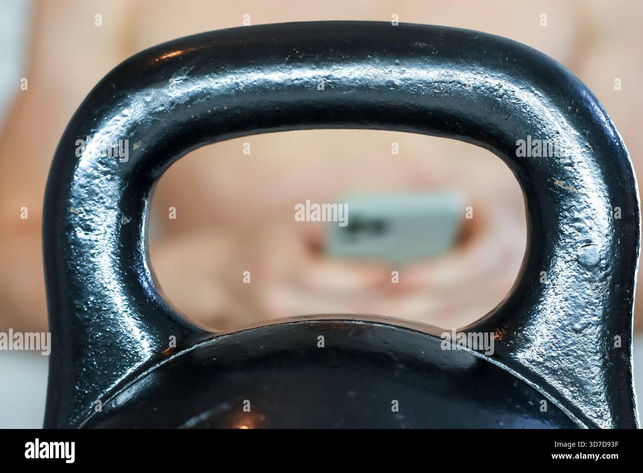Persona che tiene lo smartphone vista attraverso la maniglia kettlebell. Vista sfocata dell'atleta che controlla il telefono attraverso il manico del kettlebell in palestra Foto Stock