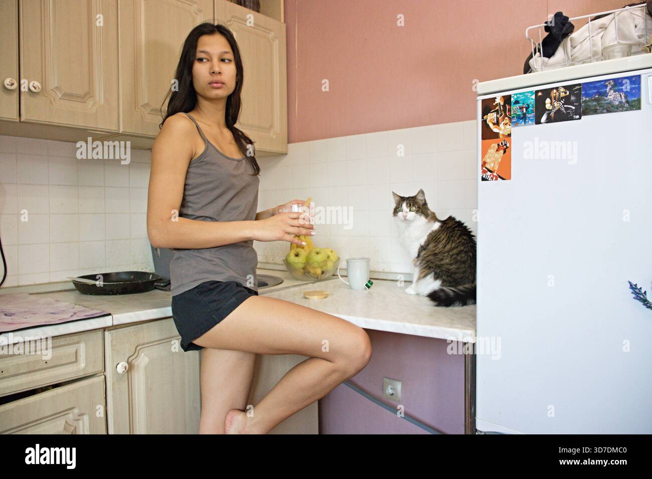 Donna indiana con canotta e pantaloncini corti siede sul bancone della cucina, reggendo un bicchiere d'acqua. Il gatto soffice siede accanto a lei vicino al frigorifero. Frutta, padella, con Foto Stock