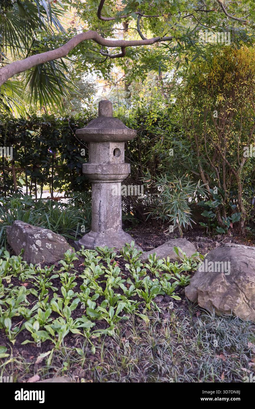 Lanterna in pietra ornata immersa in un tranquillo giardino con rami di alberi sovrastanti, fitti arbusti di fogliame, rocce aspre e ammassi di fresche piante verdi Foto Stock