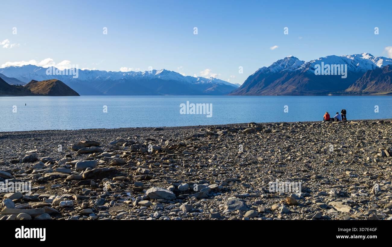 Una spiaggia rocciosa sulla riva del lago Hawea nell'Isola del Sud della nuova Zelanda. Sullo sfondo si trovano le montagne innevate delle Alpi meridionali Foto Stock