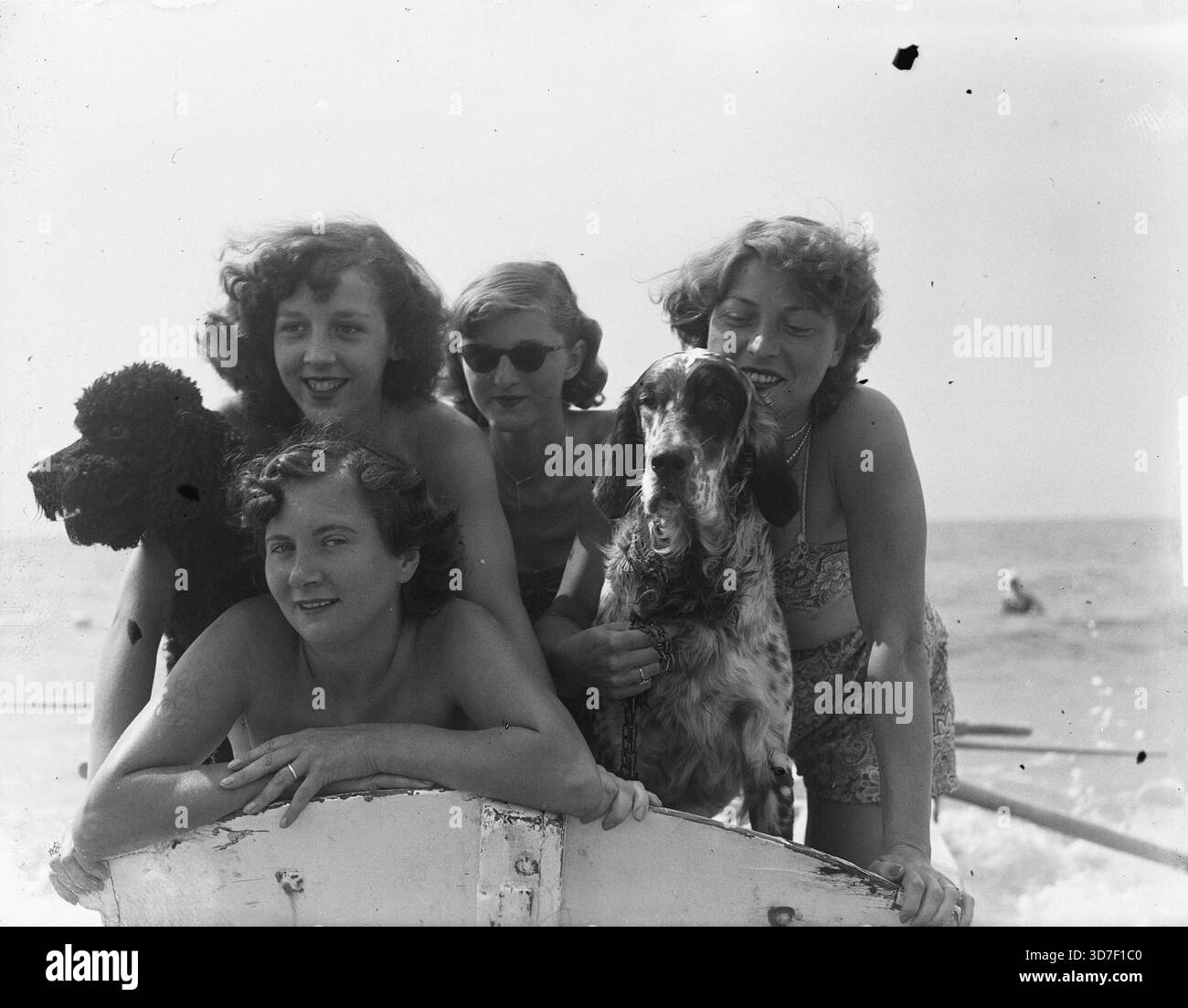 Zandvoort, Paesi Bassi, 1950 – vivace scenario di spiaggia in bianco e nero, che mostra un gruppo di giovani donne eleganti che posano gioiosamente su una barca con i loro cani in mare. I capelli baciati dal sole, i sorrisi luminosi e le giocose energie marine creano un'atmosfera estiva spensierata, mentre i cani aggiungono fascino e spontaneità. Un elegante assaggio della cultura del tempo libero del dopoguerra sulla costa olandese, piena di calore, amicizia ed eleganza da spiaggia. Foto Stock