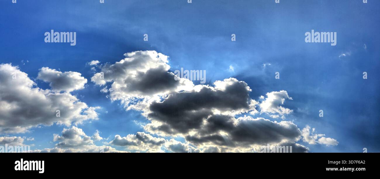 Una vista grandangolare che mostra morbide nuvole sparse contro un vasto cielo blu con la luce del sole che sbircia, creando un ambiente sereno e rinfrescante Foto Stock