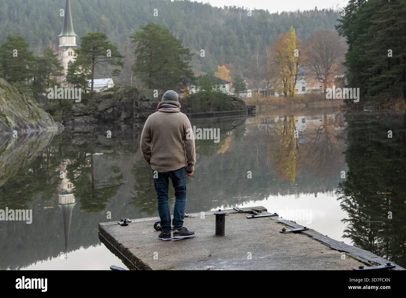 L'uomo si trova su un molo vicino a un lago calmo, circondato da vibranti alberi autunnali e da una chiesa lontana, creando un ambiente sereno e riflettente Foto Stock
