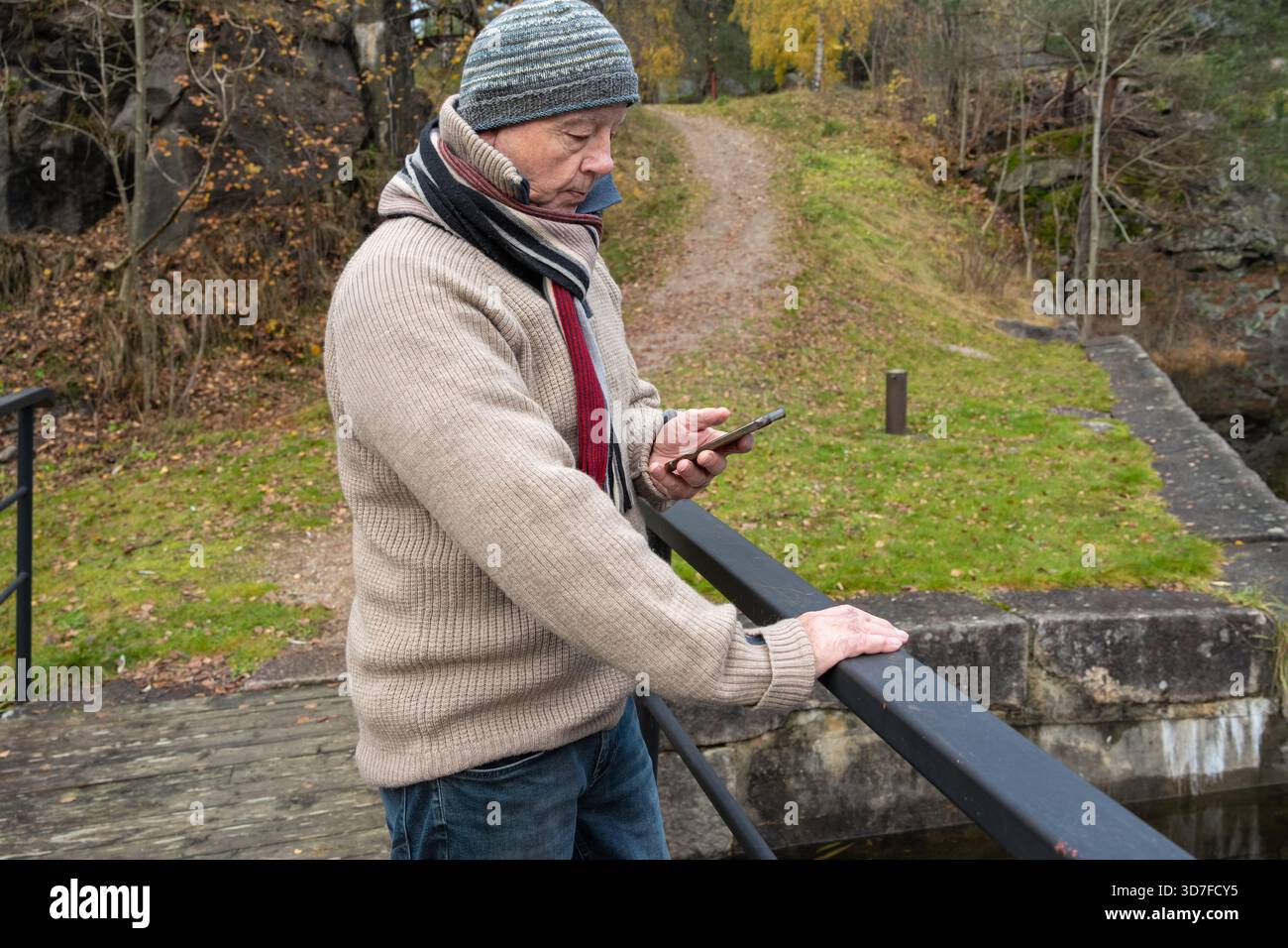 Uomo anziano in abiti caldi, in piedi accanto a una ringhiera, impegnato con lo smartphone in un ambiente all'aperto sereno con colori autunnali e una pacca naturale Foto Stock
