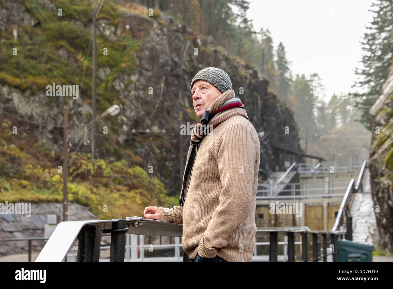 L'uomo più anziano con un maglione caldo e una sciarpa si erge accanto a una ringhiera, circondato da scogliere rocciose e alberi, apprezzando la tranquilla atmosfera all'aperto e il natu Foto Stock