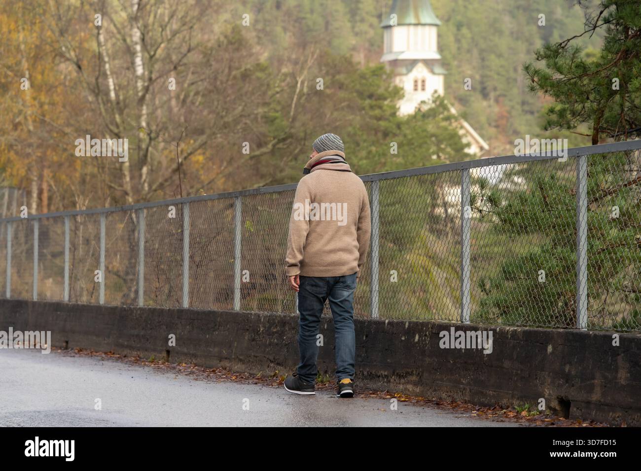 Indossando un maglione caldo, passeggia lungo una strada piovosa accanto a una recinzione, con un vivace fogliame autunnale e una chiesa sullo sfondo, creando un ambiente sereno Foto Stock