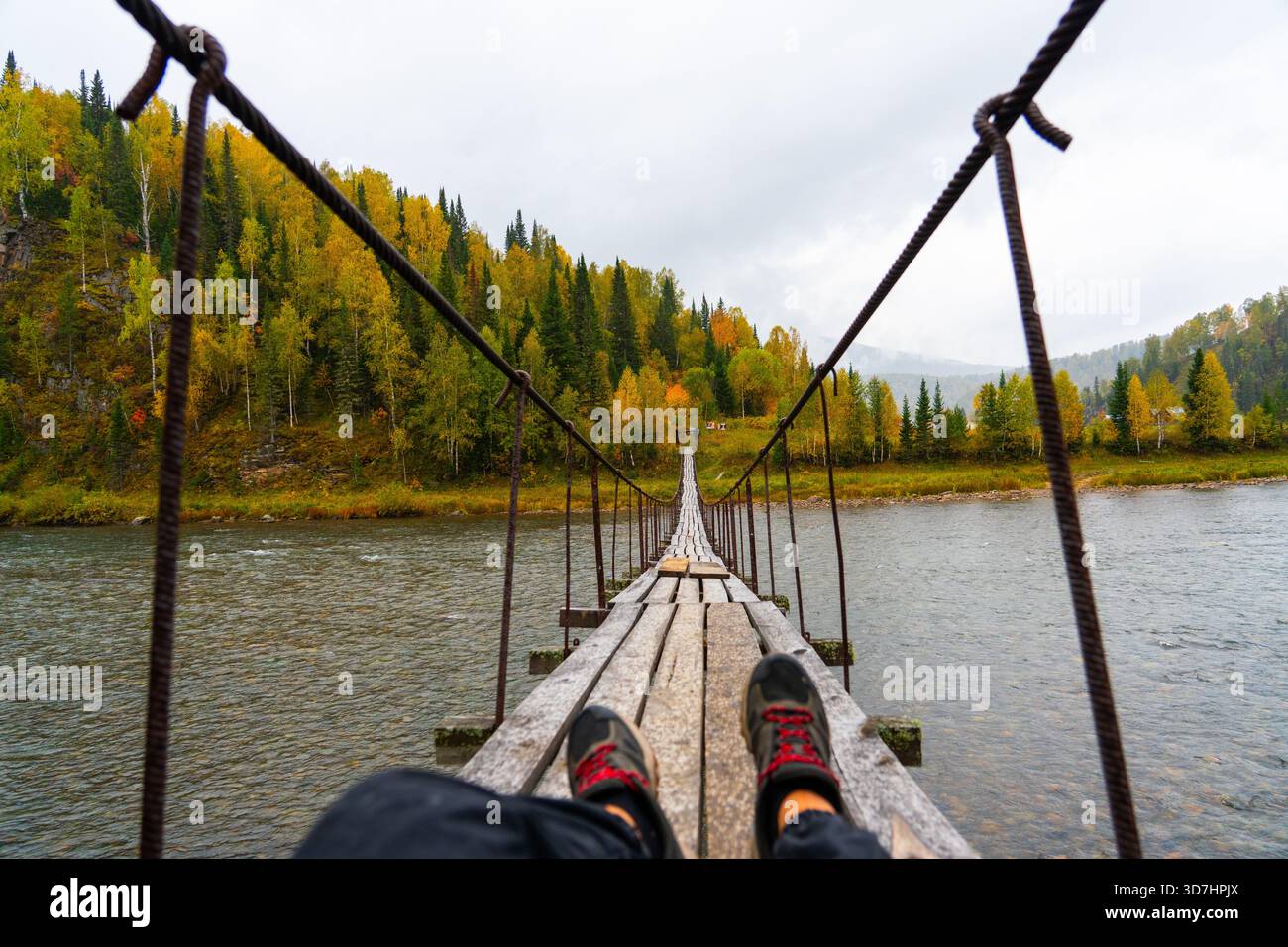 Punto di vista in prima persona di una persona seduta su un ponte sospeso in legno che guarda verso la sponda opposta coperta da una fitta foresta autunnale. Persona p Foto Stock