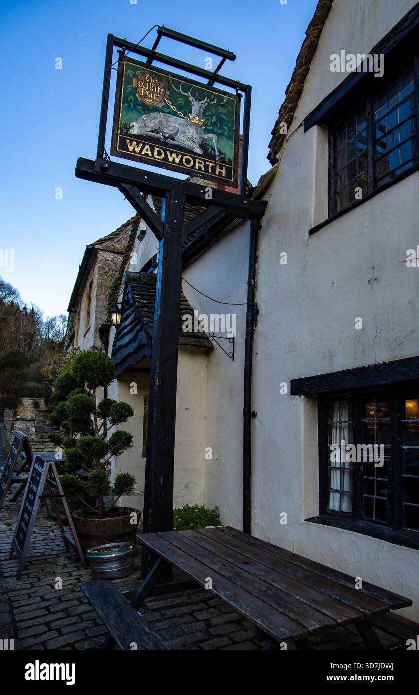 Insegna tradizionale inglese per il birrificio Wadworth, appeso fuori dallo storico pub White Hart Castle Combe, con posti a sedere all'aperto su una strada acciottolata Foto Stock