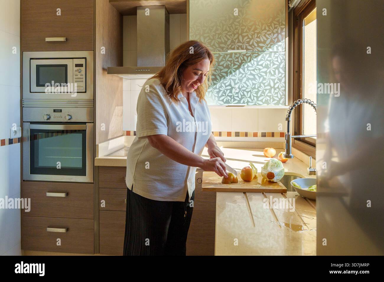 Donna felice che affetta frutta fresca su un tagliere di legno nella sua luminosa cucina, preparando un pasto nutriente e sano Foto Stock