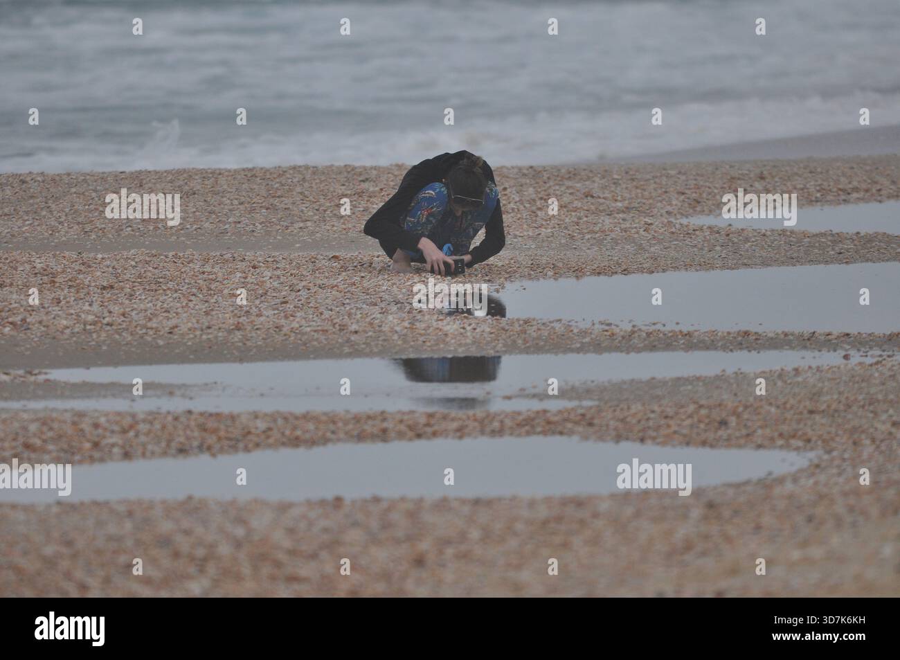Una persona accovacciata su una spiaggia di ciottoli, concentrandosi sulla cattura dei riflessi nelle piscine d'acqua poco profonde durante il tramonto. Le onde si infrangono dolcemente sulla riva come il cielo Foto Stock