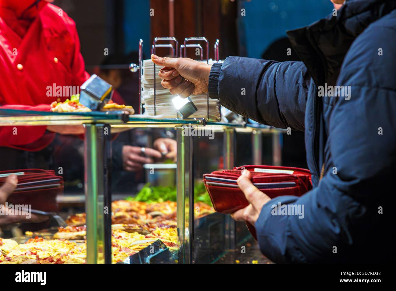 Cliente che paga per cibo di strada caldo con un portafoglio mentre dà un denaro a un venditore in una giacca rossa dietro un banco di vetro con spuntini caldi a un verricello Foto Stock