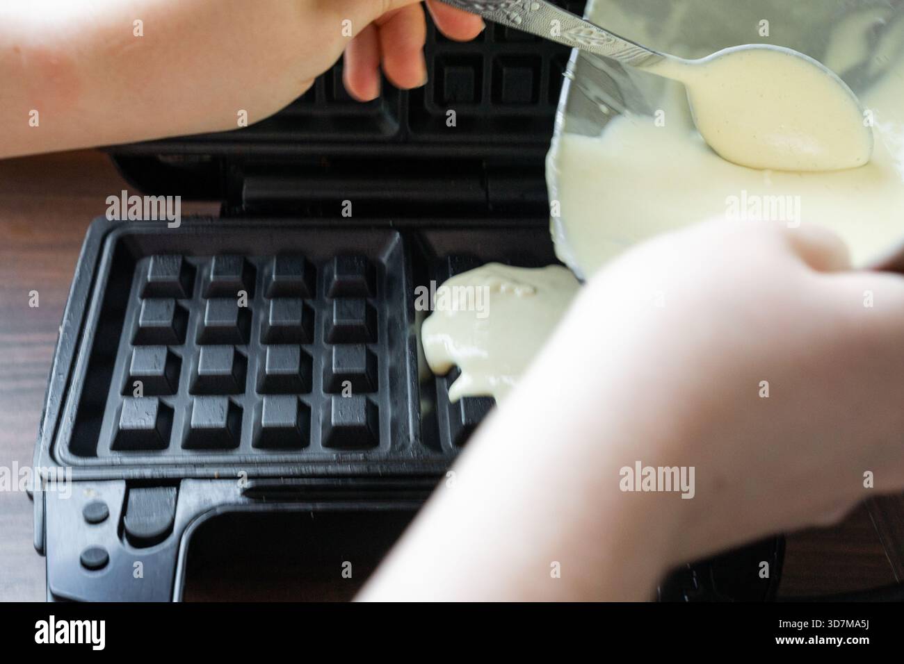 L'impasto liscio fluisce da un cucchiaio nel ferro da stiro caldo per waffle. Il momento cattura una fase iniziale della preparazione dei waffle. Foto Stock