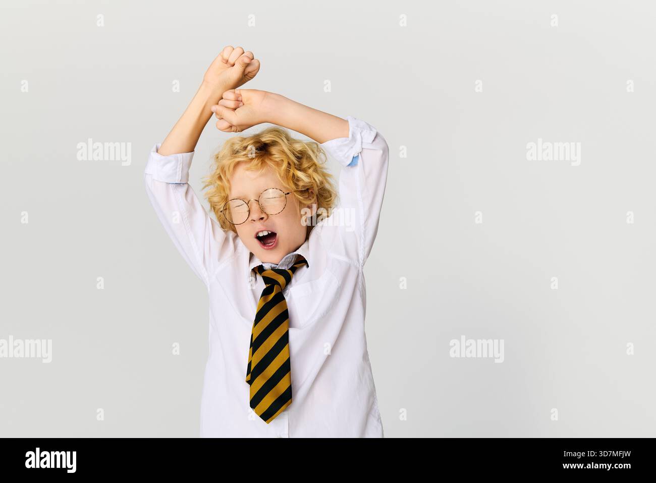 Un ragazzo felice con i capelli biondi ondulati si allunga indossando una camicia bianca e una cravatta a righe in uno studio. Foto Stock