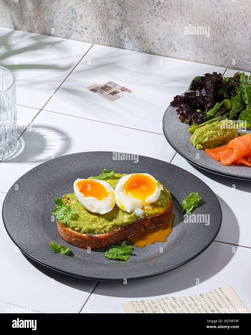 Toast all'avocado con uova sode e verdure fresche, luce del sole che proietta ombre morbide sullo sfondo delle pareti in cemento, grigio ceramica Foto Stock
