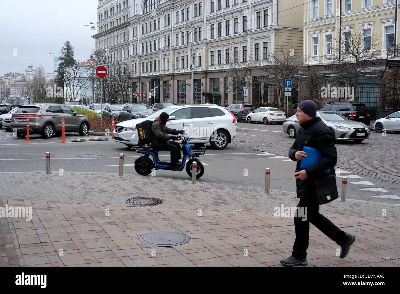 Un addetto alle consegne percorre la trafficata strada della città mentre i pedoni passano. Kiev, Ucraina, 26 novembre 2025. Foto Stock