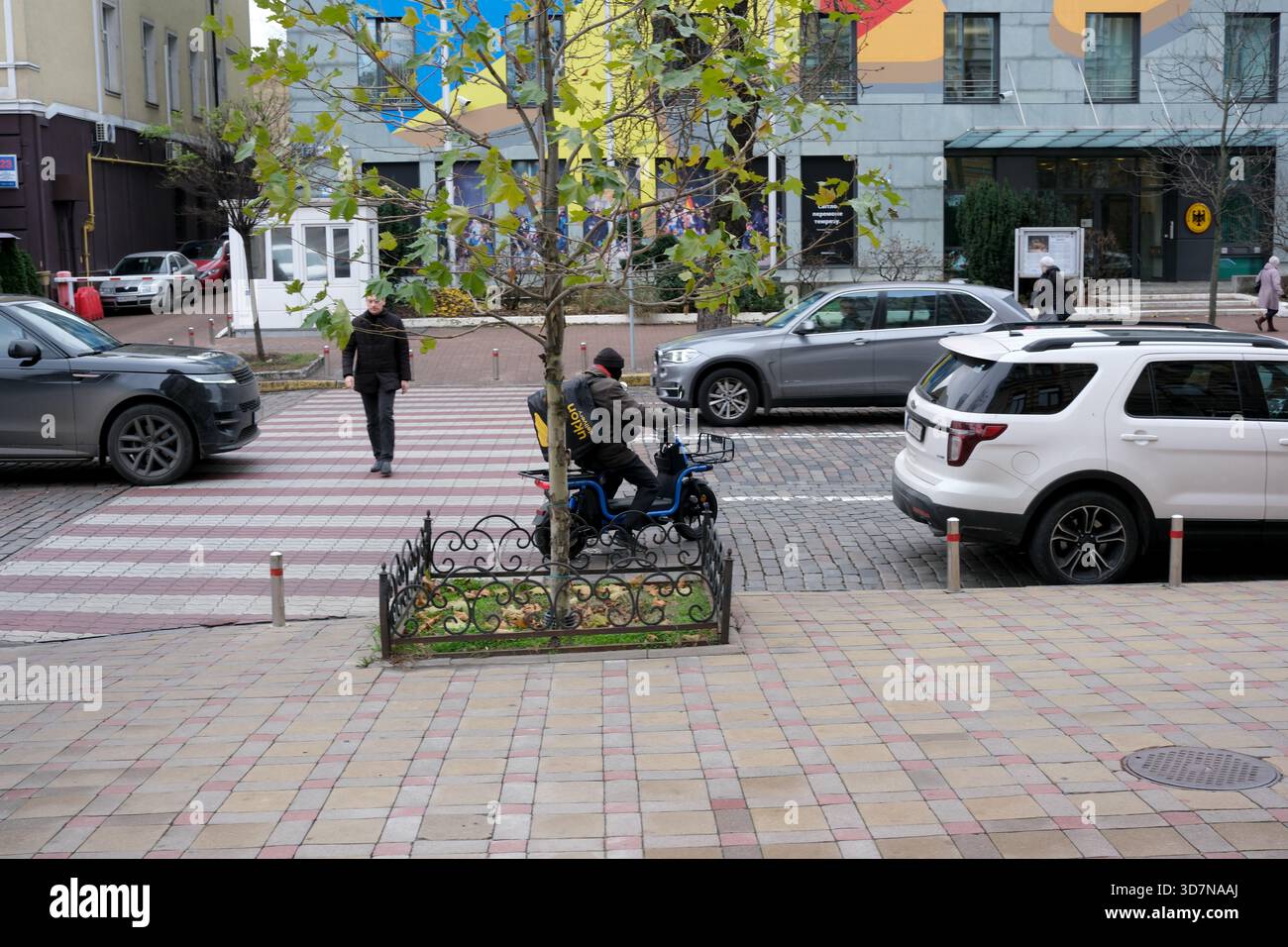 Un ciclista si prende una pausa in una tranquilla strada cittadina circondata da veicoli e pedoni. Kiev, Ucraina, 26 novembre 2025. Foto Stock