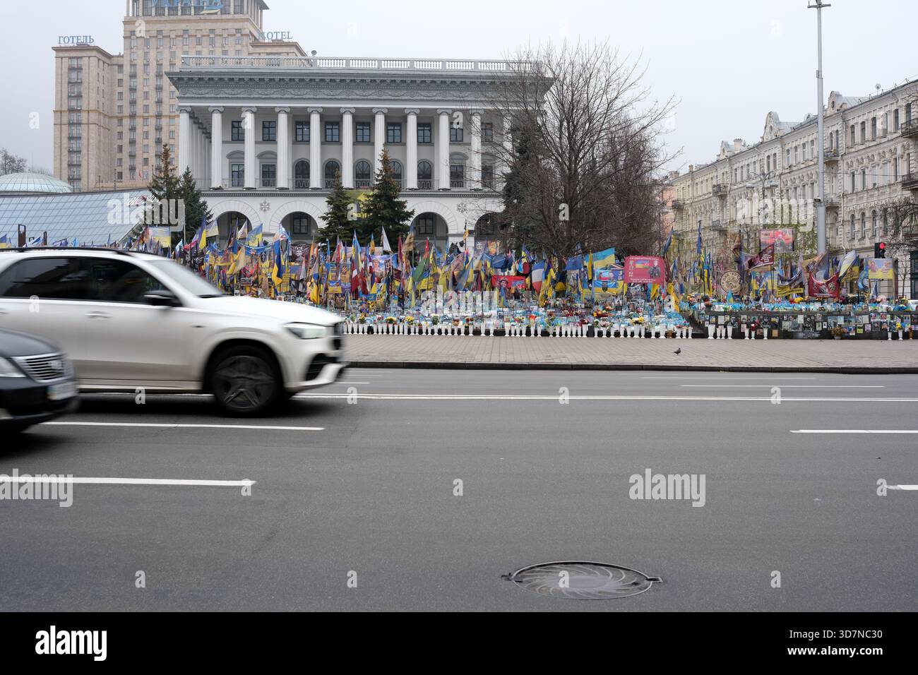 Bandiere colorate ondeggiano orgogliosamente intorno a un sito commemorativo mentre le auto passano rapidamente. Khreshchatyk, Kiev, Ucraina. 26 novembre 2025. Foto Stock