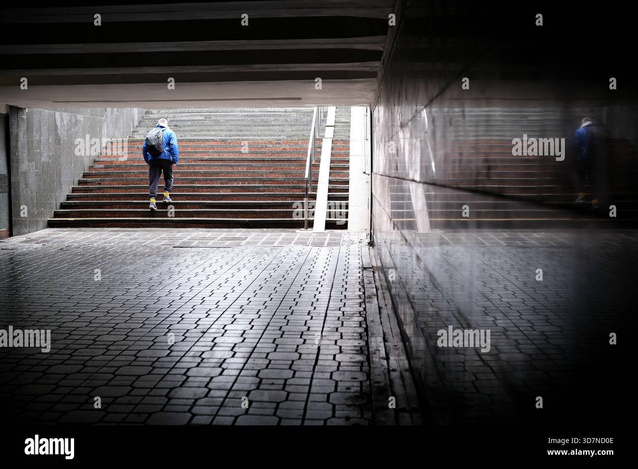 Una persona con una giacca blu cammina su scale bagnate in un tunnel urbano, riflettendo sulle pareti. Foto Stock