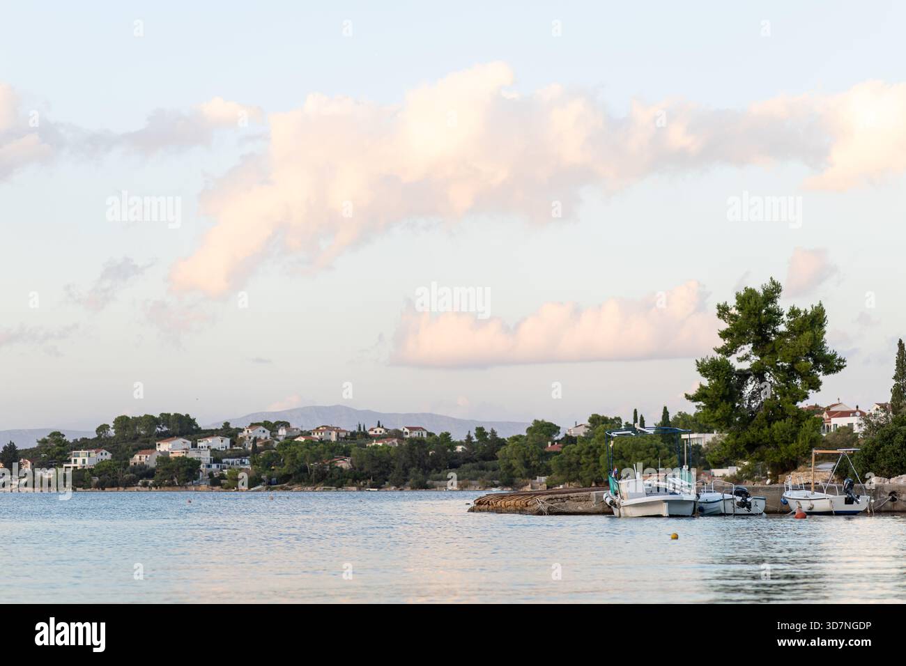 Un tranquillo villaggio sul mare lungo una baia tranquilla, con barche attraccate in un piccolo porto e case di pietra illuminate dal sole che si arrampicano su una dolce collina sotto un blu chiaro Foto Stock