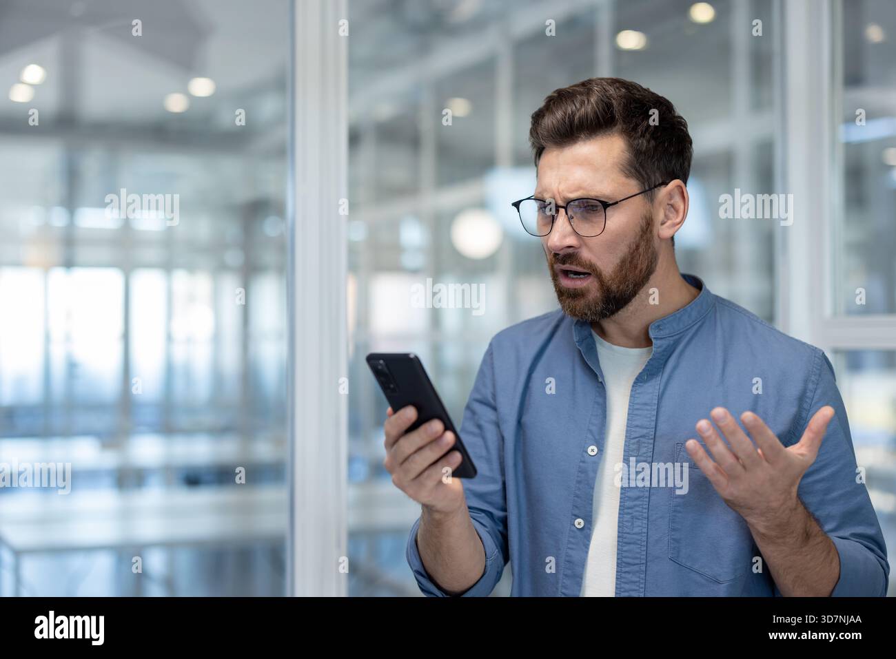 Uomo barbuto che indossa gli occhiali e una camicia blu che regge uno smartphone, esprimendo confusione e stress mentre guarda lo schermo in un moderno ambiente d'ufficio Foto Stock