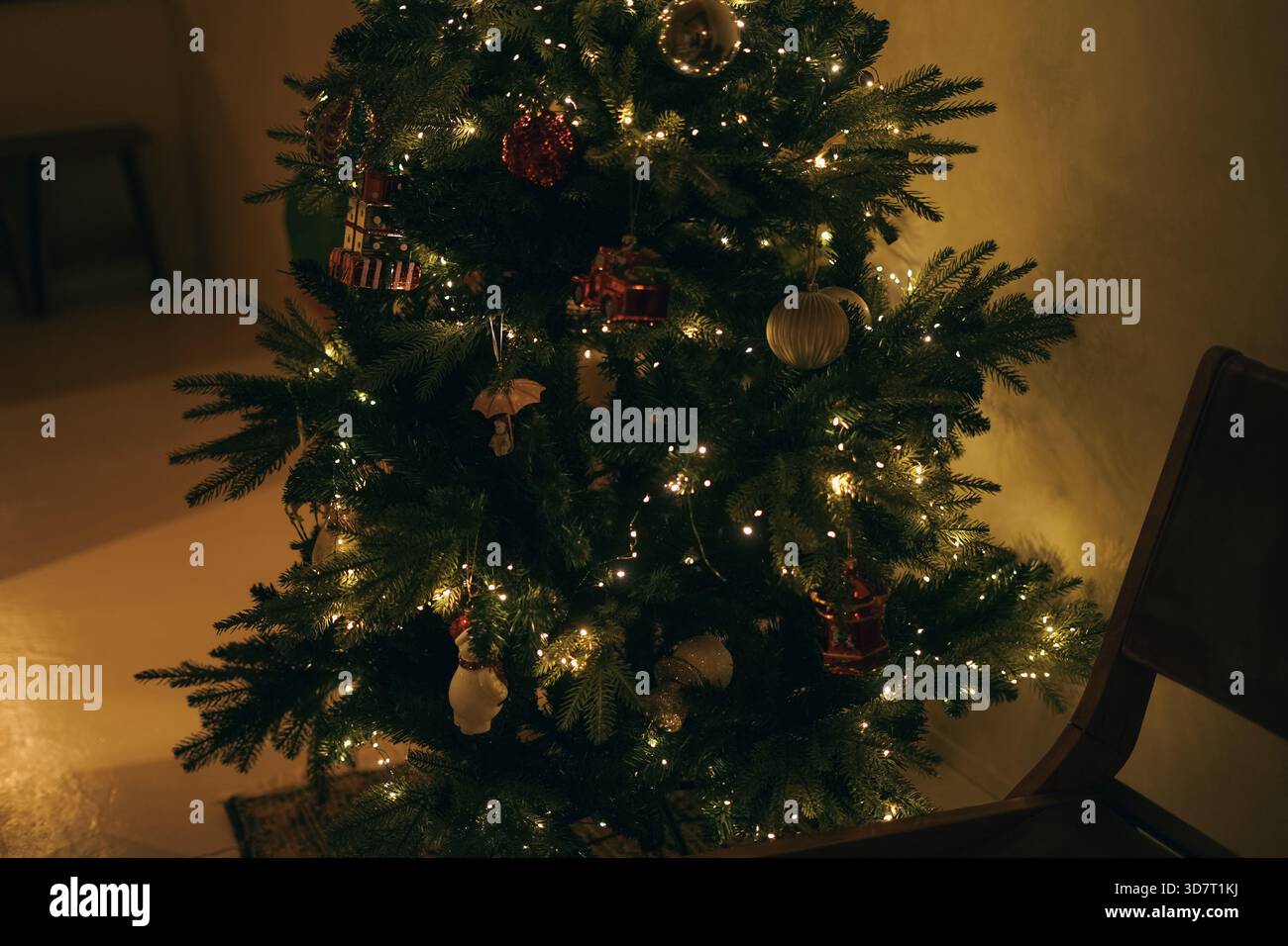 Intimo spazio interno con una sedia in legno e un albero di Natale adornato con decorazioni scintillanti sotto la luce calda Foto Stock