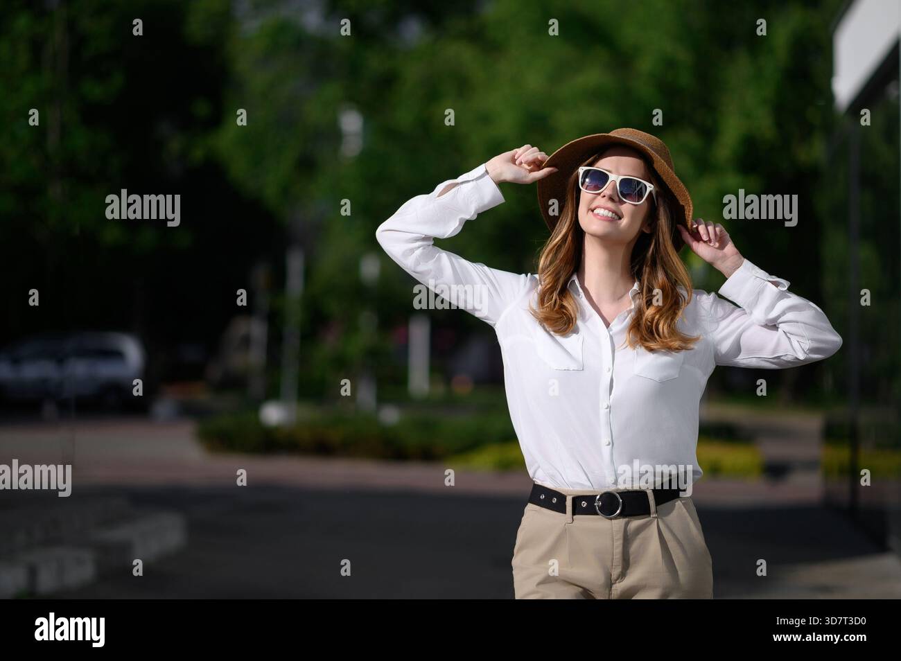 Elegante donna d'affari con occhiali da sole gode di una giornata di sole sulle strade della città. Elegante viaggiatrice di città, sofisticata donna d'affari, si aggrappa sul cappello e guarda Foto Stock