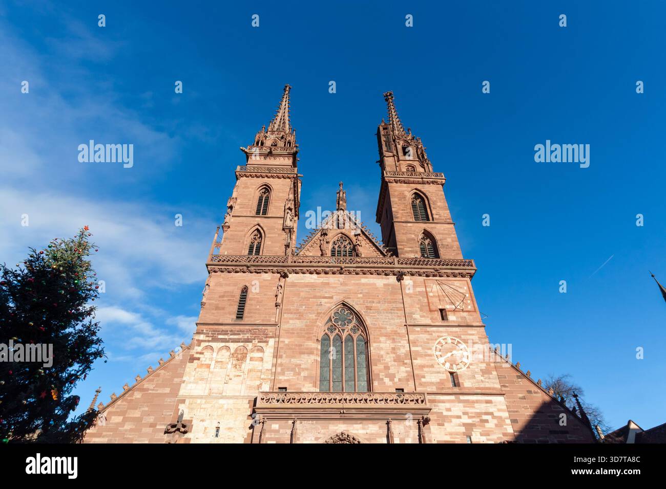Primo piano dell'impressionante facciata in arenaria rossa e delle doppie guglie gotiche della cattedrale di Basilea, con orologio e meridiana sotto un blu chiaro Foto Stock