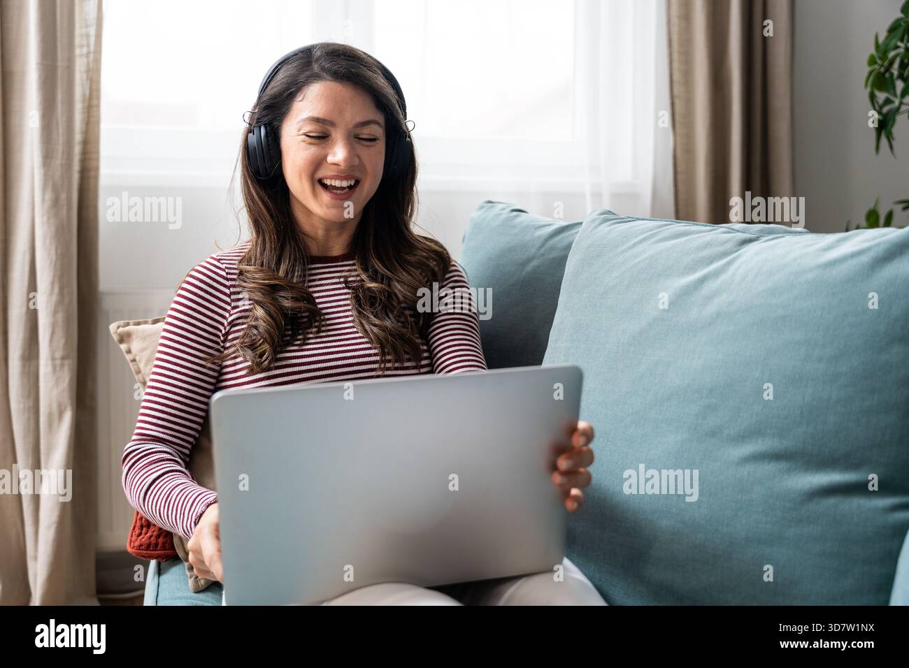Lavoro a distanza a casa, la giovane donna riproduce musica in streaming tramite le cuffie, si concentra sulla gestione di attività e messaggi online da parte di un laptop, in qualità di freelance indipendente Foto Stock
