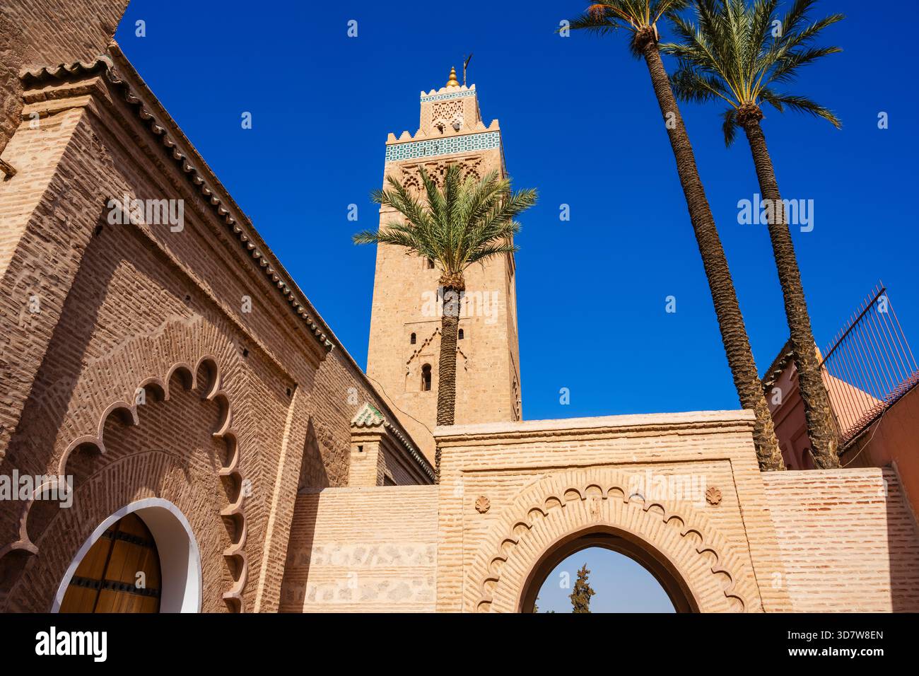 Alta torre di minareto con palme sotto un cielo blu vibrante in un ambiente architettonico storico. Marrakech, Marocco Foto Stock