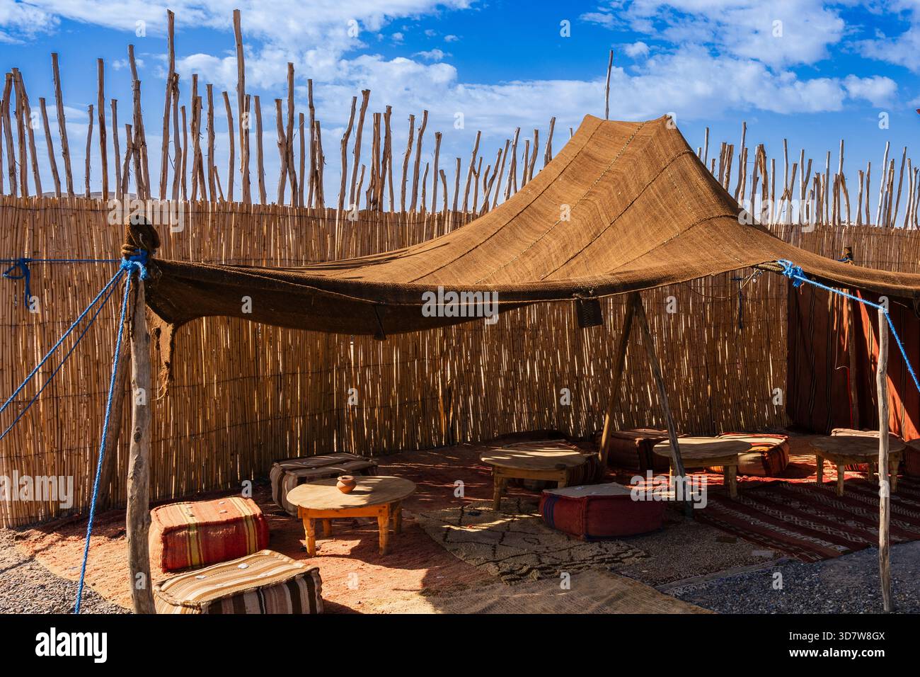 Tradizionale tenda esterna con pareti intrecciate, tavoli in legno e cuscini vibranti sotto un cielo blu. Deserto di Agafay, Marrakech-Safi, Marocco Foto Stock