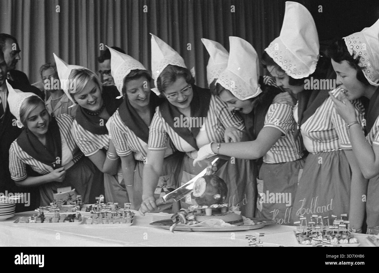 📍 Gouda, Paesi Bassi, 1961 – Un vivace gruppo di tradizionali "kaasmeisjes" olandesi (ragazze del formaggio), vestiti con camicette a righe, grembiuli e classici cappellini in pizzo, si riuniscono intorno a una torta festosa a forma di formaggio Gouda e Edam. Il loro allegro lavoro di squadra e sorrisi curiosi catturano un gioioso momento promozionale che celebra la cultura casearia olandese. Foto Stock