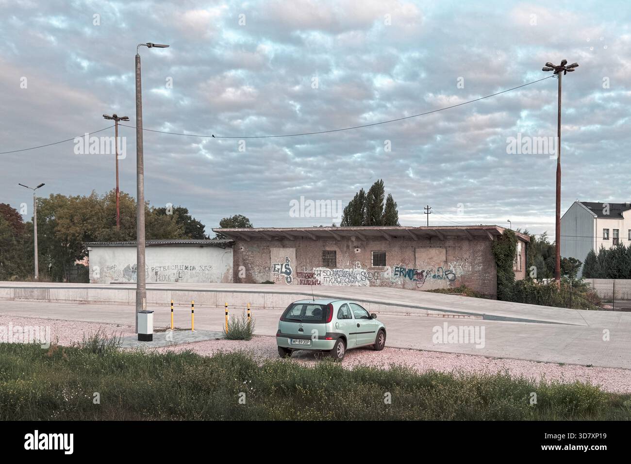 Vecchio edificio industriale coperto di graffiti con una piccola auto parcheggiata in un posto tranquillo e vuoto sotto un cielo nuvoloso. Foto Stock