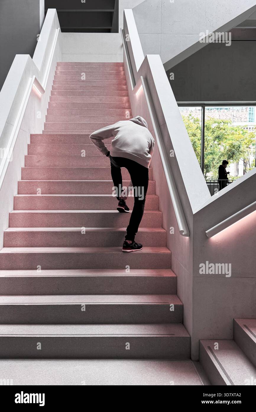 Persona che regola la scarpa stando in piedi su una scala moderna illuminata da luci rosa tenui all'interno di un edificio contemporaneo. Foto Stock