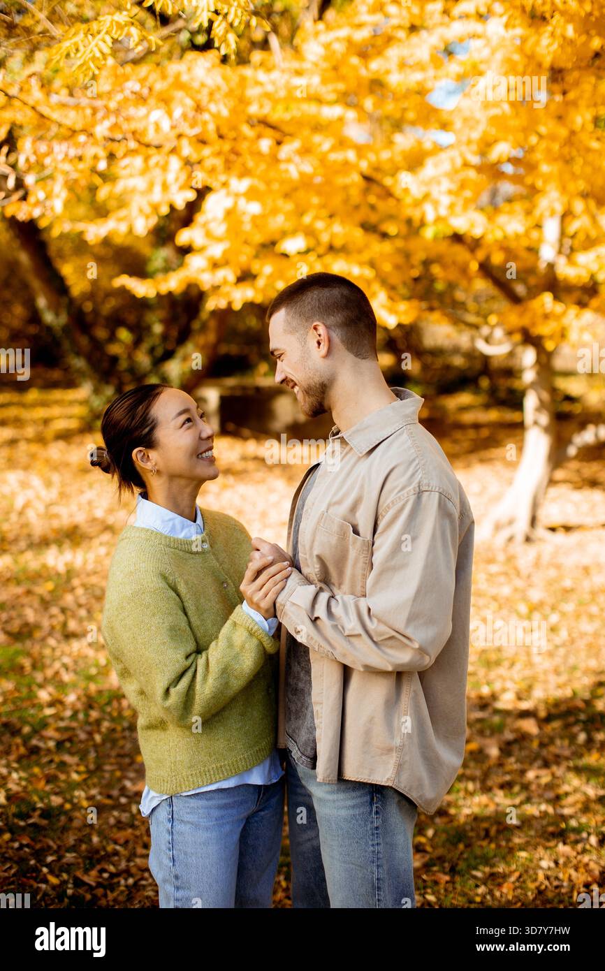 Una donna coreana e un uomo caucasico si sorridono, tenendosi per mano tra le vivaci foglie autunnali e il caldo sole. Foto Stock