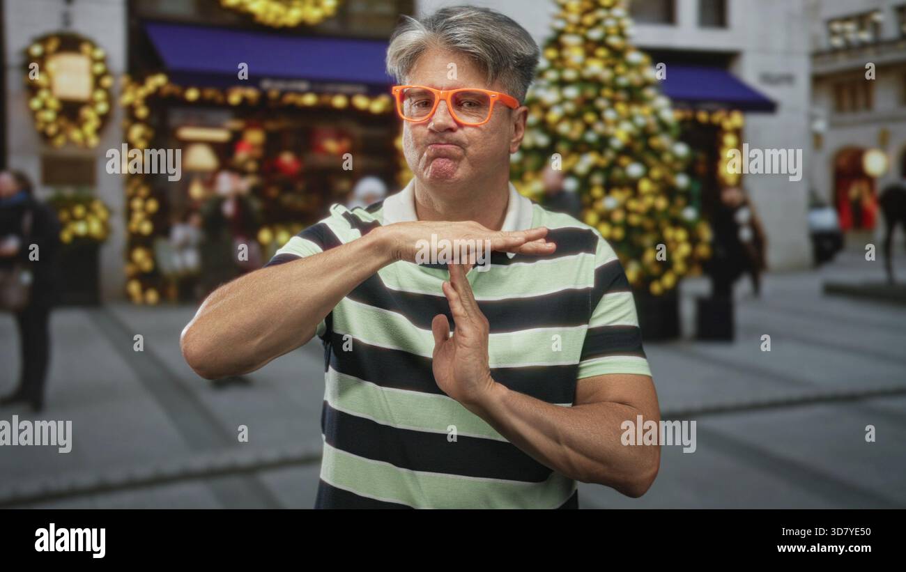 Uomo con bicchieri arancioni e polo a righe, mani che formano un cartello timeout nella strada dello shopping decorata con albero di natale e ghirlande; frustrazione. Foto Stock
