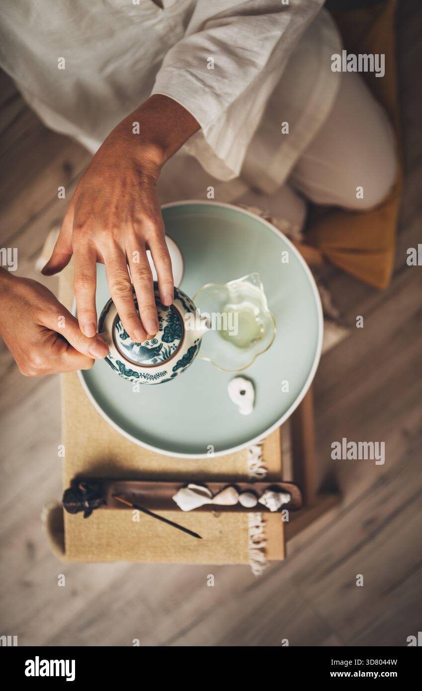 Mani maschili che versano il tè dalla teiera di argilla fantasia in una caraffa di vetro trasparente, creando un sereno rituale del tè. Luce soffusa, vassoio da tè in legno e vegetazione evocano Foto Stock