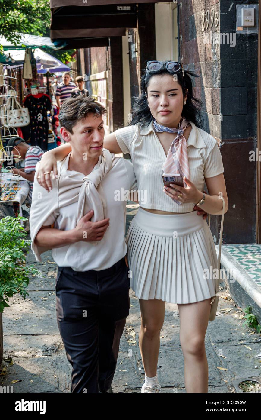 Città del Messico, Messico, Coyoacan Avenida Miguel Hidalgo, giovane coppia adulta che cammina, uomo più corto braccio intorno a una donna più alta, con in mano un negozio di smartphone sul marciapiede Foto Stock