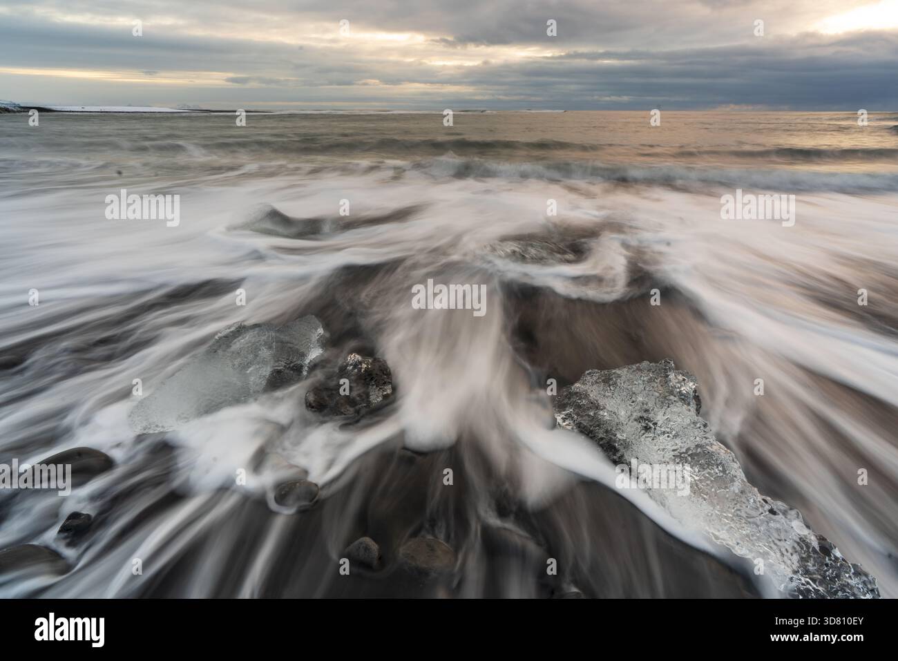 Vista dei frammenti di ghiaccio glaciali sparsi sulla spiaggia di sabbia nera, in contrasto con le onde bianche spumeggianti sotto un cielo moody, Diamond Beach, Sveitarfelag Foto Stock
