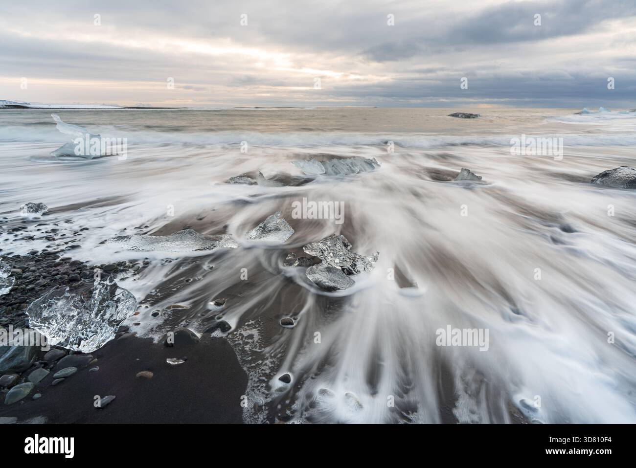 Vista dei frammenti di ghiaccio traslucidi sparsi sulle sabbie vulcaniche nere, baciati dall'etereo abbraccio delle onde schiumose sotto un cielo sereno, Diamond Foto Stock