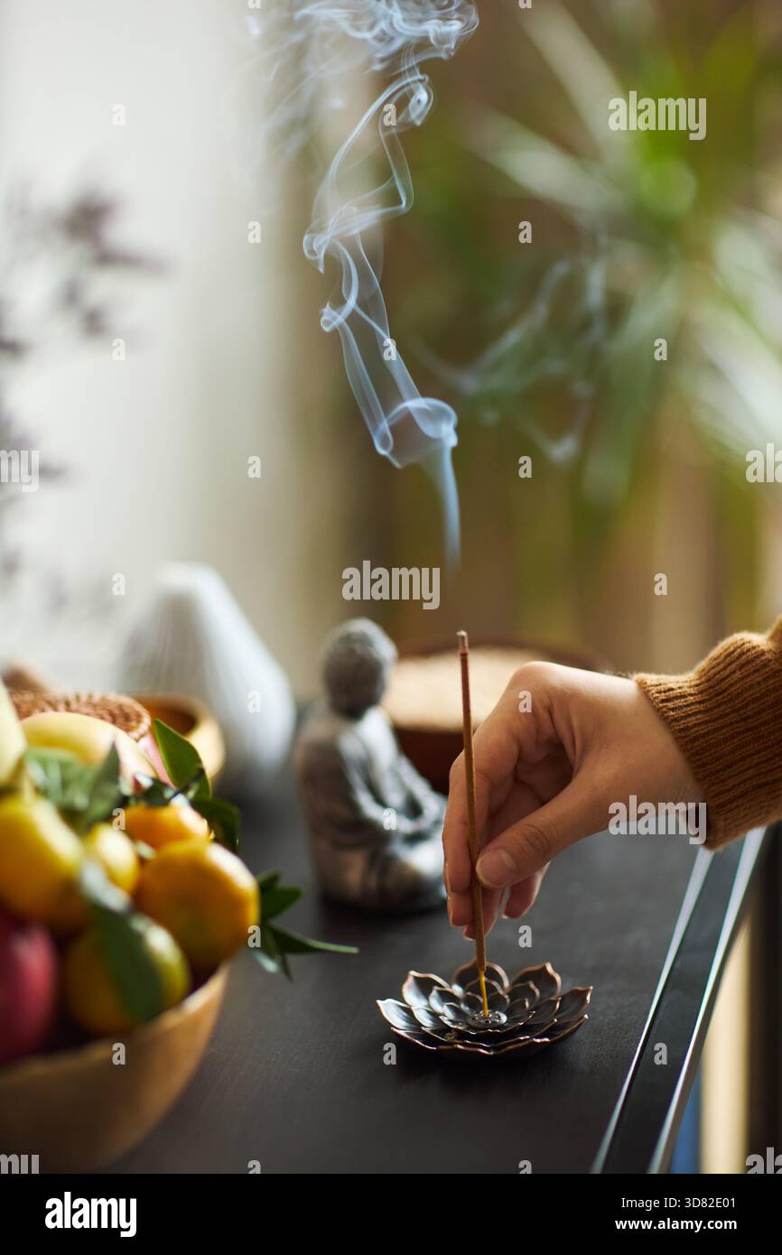 Giovane donna asiatica adulta che accende a mano il bastoncino di incenso in un supporto a forma di loto con lievitazione di fumo, cesto di frutta e piccola statua di Buddha sul tavolo, ambiente interno con piante sfocate Foto Stock