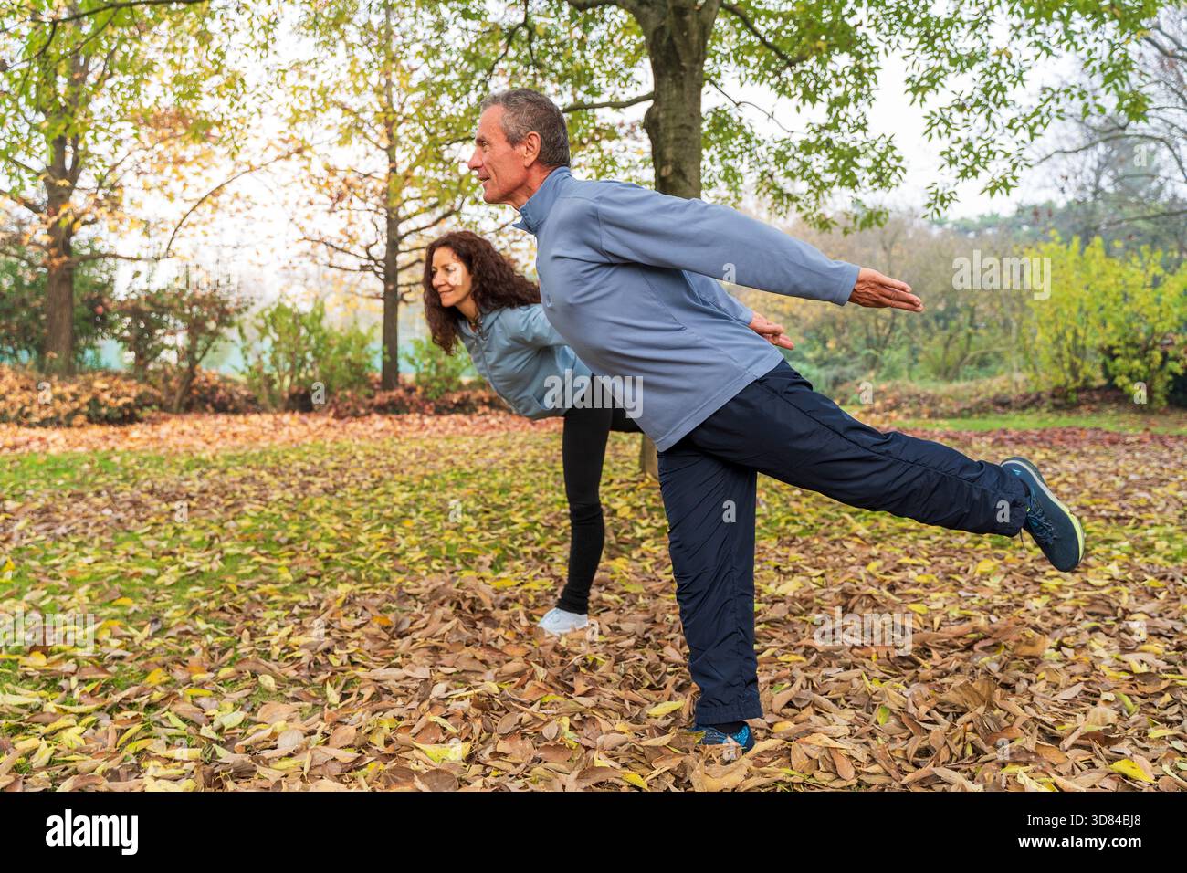 Coppia matura in equilibrio su una gamba, praticando tai chi in un parco autunnale con foglie cadute Foto Stock