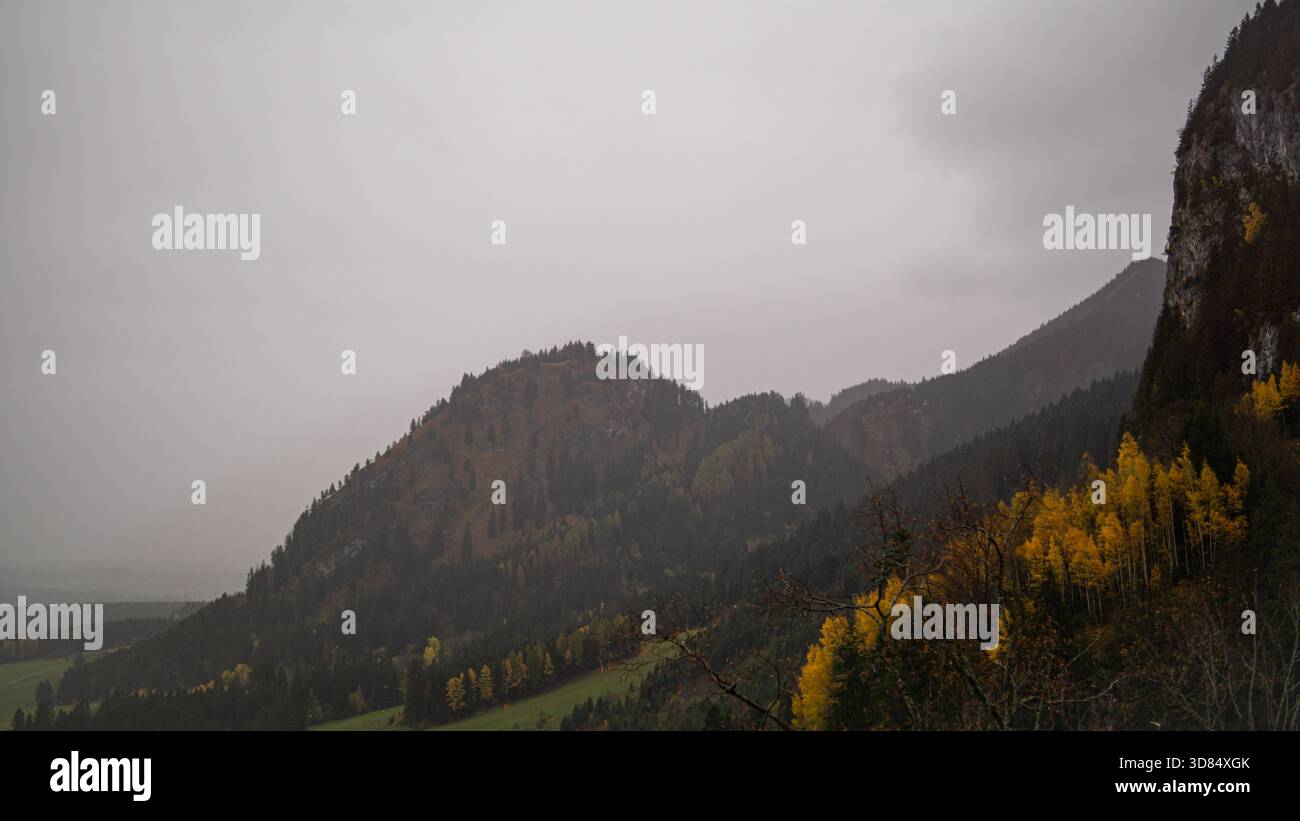 Una spettacolare montagna nebbiosa sorge sopra colline boscose punteggiate da alberi autunnali gialli. Foto Stock