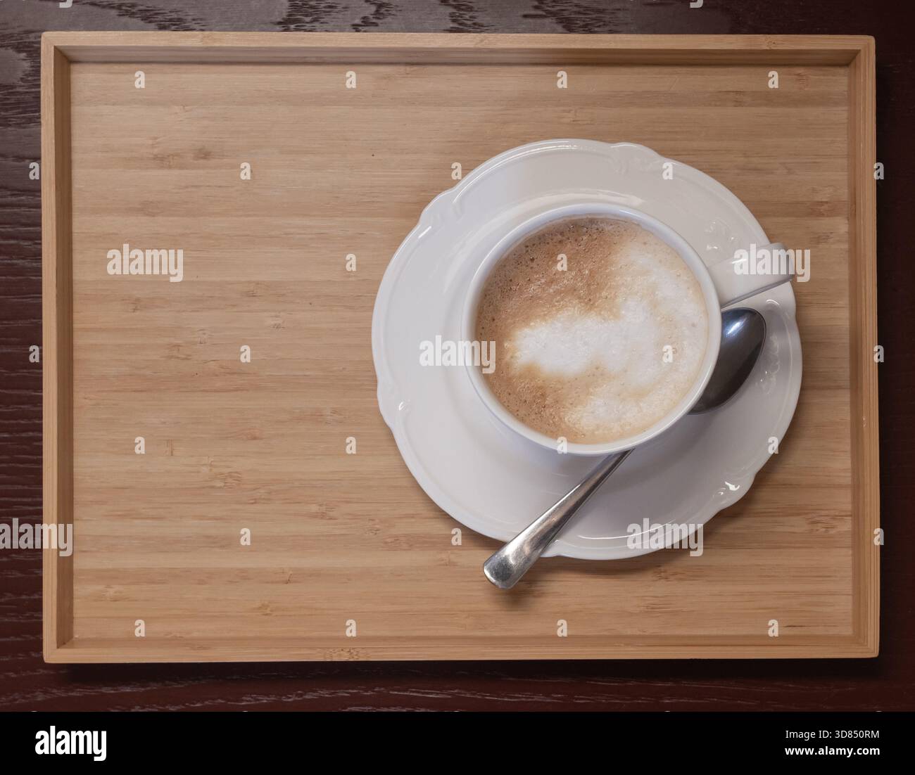Cappuccino con schiuma cremosa servito in una tazza bianca su un vassoio di legno con cucchiaio. Ambiente accogliente e caldo per caffe', colazione e temi di stile di vita. Foto Stock