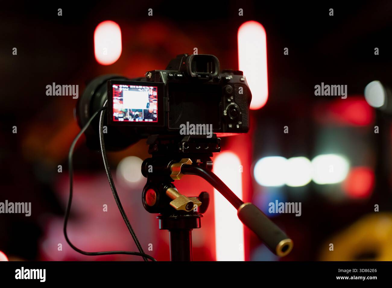 Vista ravvicinata della telecamera montata su un cavalletto video con impugnatura visibile, testa regolabile e display illuminato su uno sfondo rosso leggermente sfocato Foto Stock