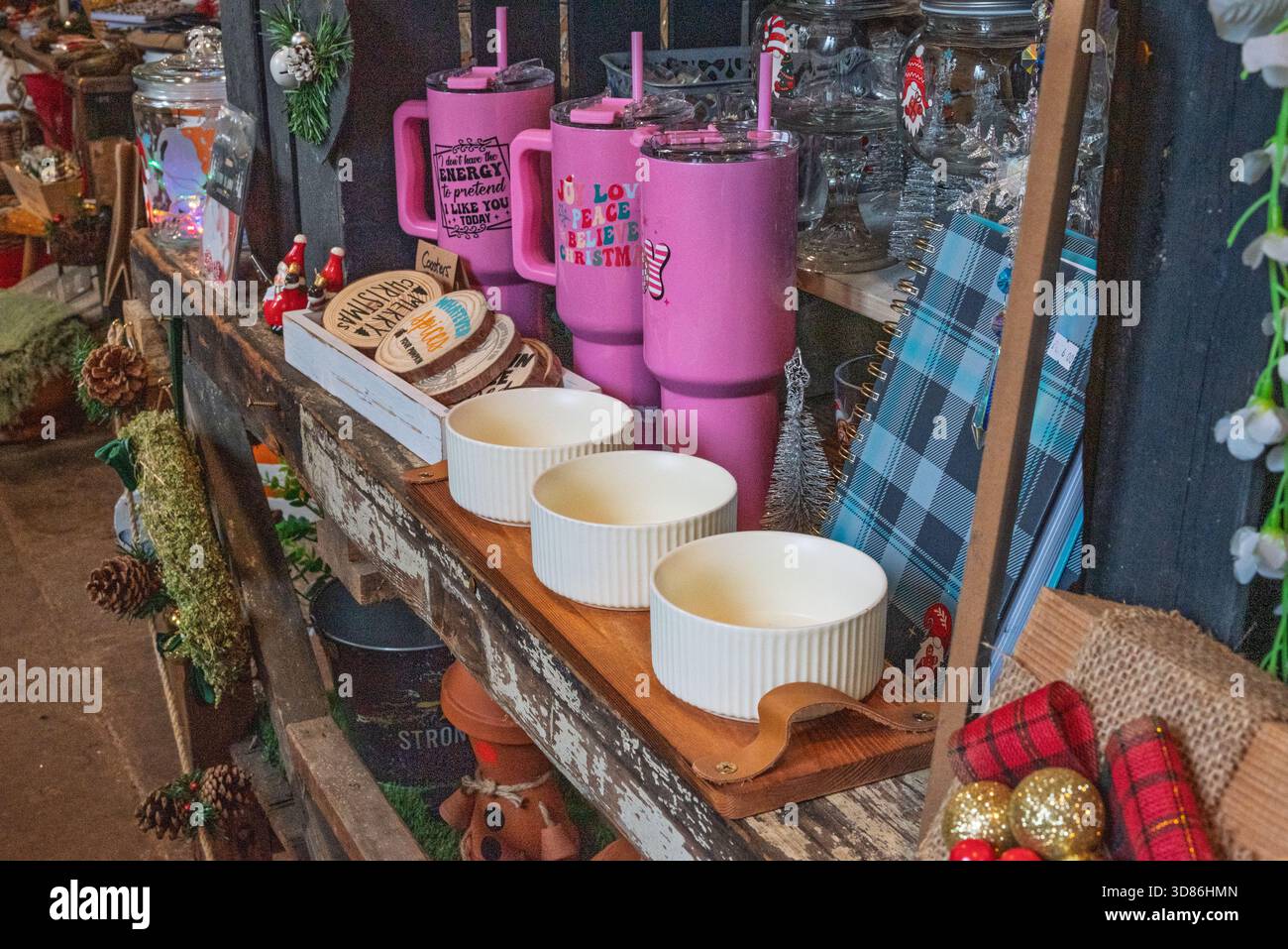 Un'esposizione festiva di oggetti a tema natalizio, tra cui bicchieri rosa, sottobicchieri in legno e pineconi decorativi, disposti su scaffali rustici in legno. Foto Stock