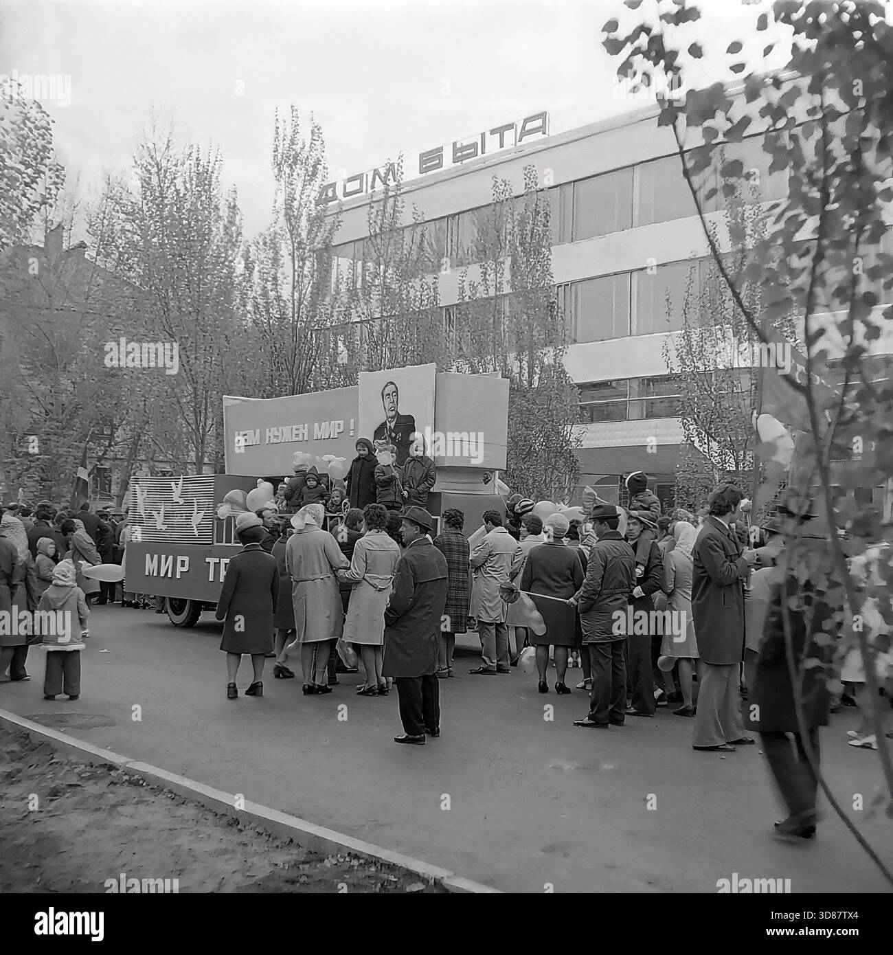 Slavyansk, Ucraina, URSS - 1970s: Una sfilata di manifestazioni festive in via Karl Marx (ora via Tsentralna). Le colonne dei residenti marciano oltre l'edificio modernista "Dom Byta" (House of Life Services). I partecipanti portano bandiere, palloncini, striscioni con slogan come "abbiamo bisogno di pace!" e ritratti di leader sovietici, tra cui Leonid Brezhnev. La gente è vestita con cappotti autunnali. Questa foto d'archivio in bianco e nero cattura i rituali ideologici, la moda e l'atmosfera sociale della tranquilla città industriale di Donbas. Foto Stock