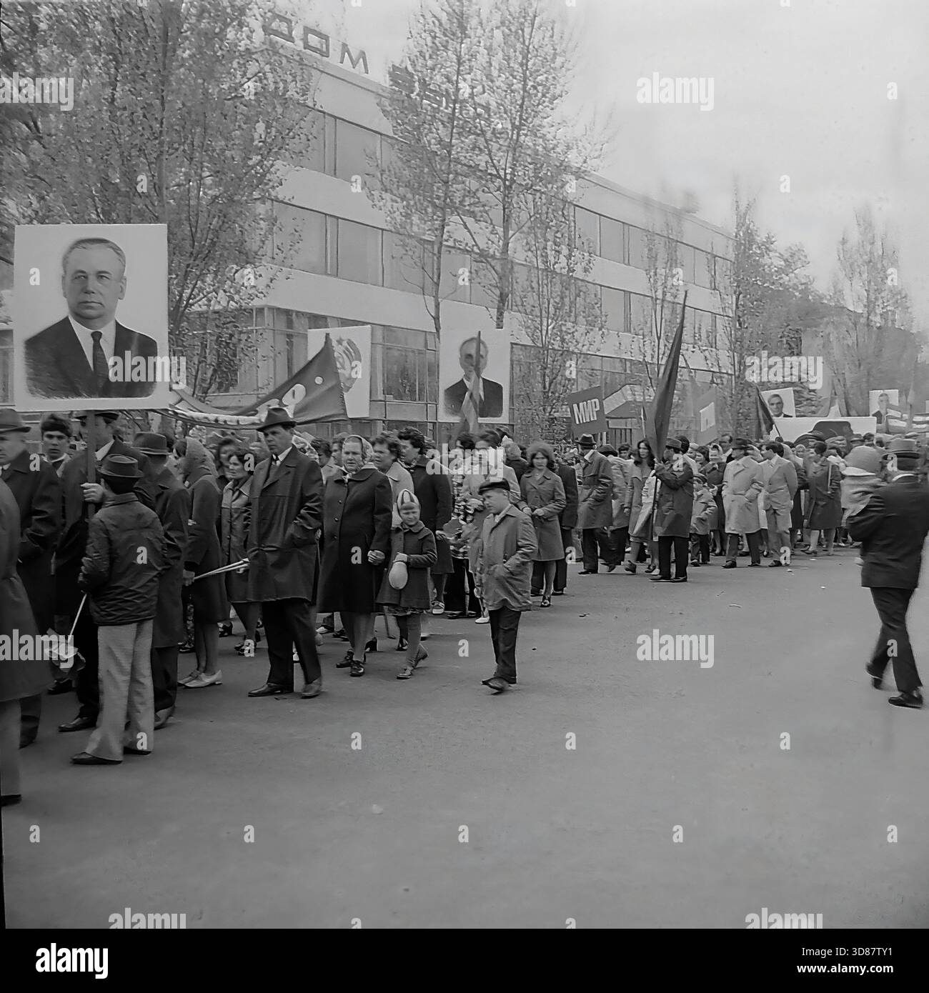 Slavyansk, Ucraina, URSS - 1970s: Una sfilata di manifestazioni festive in via Karl Marx (ora via Tsentralna). Le colonne dei residenti marciano oltre l'edificio modernista "Dom Byta" (House of Life Services). I partecipanti portano bandiere, palloncini, striscioni con slogan come "abbiamo bisogno di pace!" e ritratti di leader sovietici, tra cui Leonid Brezhnev. La gente è vestita con cappotti autunnali. Questa foto d'archivio in bianco e nero cattura i rituali ideologici, la moda e l'atmosfera sociale della tranquilla città industriale di Donbas. Foto Stock