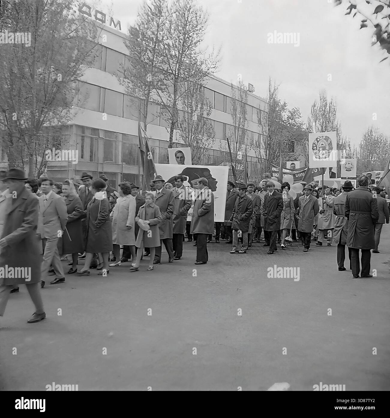 Slavyansk, Ucraina, URSS - 1970s: Una sfilata di manifestazioni festive in via Karl Marx (ora via Tsentralna). Le colonne dei residenti marciano oltre l'edificio modernista "Dom Byta" (House of Life Services). I partecipanti portano bandiere, palloncini, striscioni con slogan come "abbiamo bisogno di pace!" e ritratti di leader sovietici, tra cui Leonid Brezhnev. La gente è vestita con cappotti autunnali. Questa foto d'archivio in bianco e nero cattura i rituali ideologici, la moda e l'atmosfera sociale della tranquilla città industriale di Donbas. Foto Stock